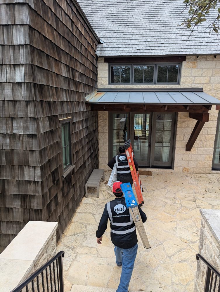 Two Ned Air Filters technicians in branded safety vests carrying ladders and new AC filters to a home in the Texas Hill Country